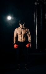 Fototapeta na wymiar Young athletic boxer in red gloves and black sportswear standing next to a punching bag. Handsome athlete showing muscles and half-naked torso. Concept of strentgh and motivation