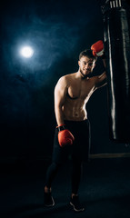 Obraz na płótnie Canvas Smiling handsome boxer in red gloves and black sportswear leans against a punching bag and poses for camera. Half-naked man's torso covered in sweat. Concept of strentgh and motivation