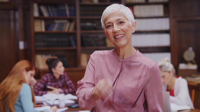 Portrait of mature professor with crossed arms standing in university library and looking at camera with copy space. Happy senior woman at the library working as a librarian. Satisfied college teacher