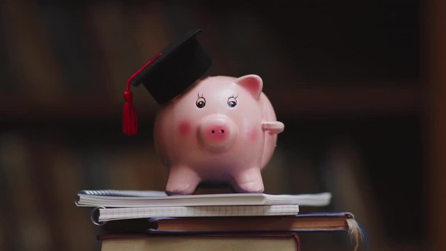 Pink piggy bank wearing graduation cap and holding pencil on pile of books in college library. Student loan and debt concept. Close up of piggybank with black graduate hat.