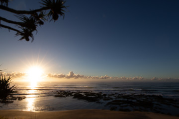 Fototapeta na wymiar Sun rise at the beach of village of Yeppoon, Queensland, Australia. At the shore of the pacific ocean near Sunshine Coast. Sun rising in between the clouds and reflecting in the water of the sea. 