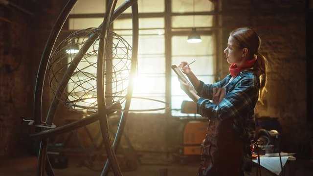 Beautiful Female Artist Sketches on a Tablet Computer Next to Brutal Metal Sculpture in Studio. Tomboy Girl Wears Checkered Shirt and Apron. Contemporary Fabricator Creating Abstract Steel Art.