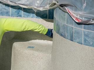Man using a plastic trowel to smooth and shape newly applied textured cement containing pebbles on a swimming pool.