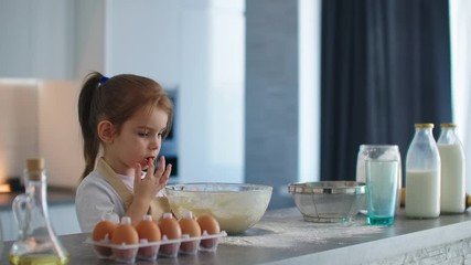 A little girl licks her finger standing in the kitchen in an apron after cooking dough with her mother