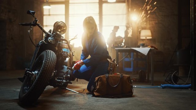 Young Beautiful Female Mechanic is Working on a Custom Bobber Motorcycle. Talented Girl Wearing a Blue Jumpsuit. She Uses a Ratchet Spanner to Tighten Nut Bolts. Creative Authentic Workshop Garage.