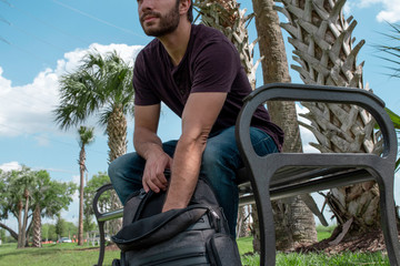 A man wearing a red shirt and blue jeans stretches his arm out reaching down into a black backpack searching on a bright sunny day on a park bench in front of some palm trees surrounded by green grass