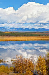Calm autumn landscape with a mirror reflection of clouds in the blue water of a pond and mountain peaks in the distance. Siberia, Buryatia, Eastern Sayan Mountains, Tunka Valley