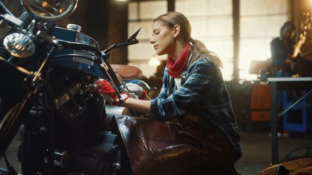 Young Beautiful Female Mechanic is Working on a Custom Bobber Motorcycle. Talented Girl Wearing a Checkered Shirt and an Apron. She Turns and Smiles at the Camera. Creative Authentic Workshop Garage.