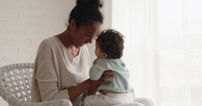 Loving mixed race mom playing lifting cute baby daughter sitting on chair. Happy family young adult african mother having fun with adorable infant child. Mum babysitter and small kid cuddling at home