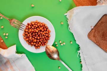 white plate with ready-baked chickpeas on a cotton vintage napkin and an old metal fork and spoon on a clean green background with scattered grains of raw chickpeas. Content for healthy eating.