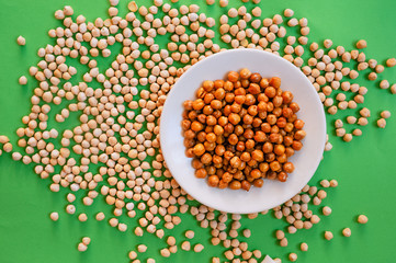 A white porcelain plate with ready-baked chickpeas stands on light raw chickpeas on a green background.