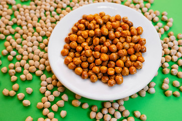 A white porcelain plate with ready-baked chickpeas stands on light raw chickpeas on a green background.