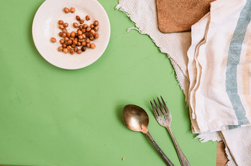 white plate with ready-baked chickpeas on a cotton vintage napkin and an old metal fork and spoon on a clean green background with scattered grains of raw chickpeas. Content for healthy eating.