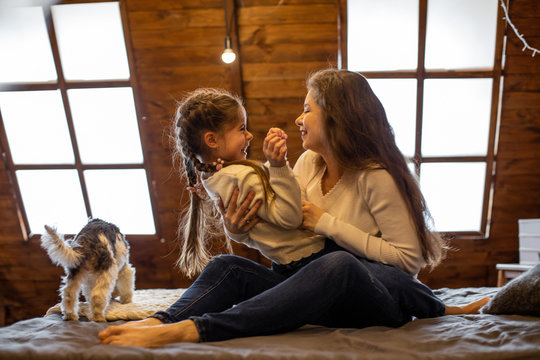 horizontal photo of a laughing girl and her long-haired mom sitting on a bed in casual clothes and their funny dog
