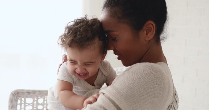 Funny adorable healthy african american baby girl or boy playing with mom at home. Young loving mother hugging taking care of little infant child daughter, cuddling, enjoying sweet motherhood concept.