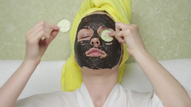 A girl with black mask on her face removes a circle of cucumber from her eyes and eats it. Charcoal face mask or black clay mud. Natural cosmetic concept. Skin care, acne treatment, cleansing skin.
