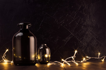 Christmas cozy dark modern interior - back and golden decoration - twinkle warm garland with elegant bottles on dark wood table and  plaster wall.