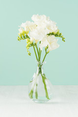 Romantic spring flowers freesia in transparent glass vase in green mint menthe interior on white wood board, vertical.