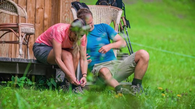 An Older Man And A Blonde Woman Who Is Tying Her Shoelace, Getting Ready For Their Hiking Trip In A Green Valley.