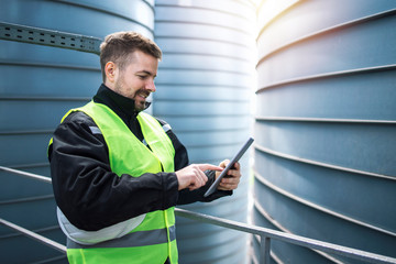 Factory worker using tablet computer in industrial factory building. In background silo storage...