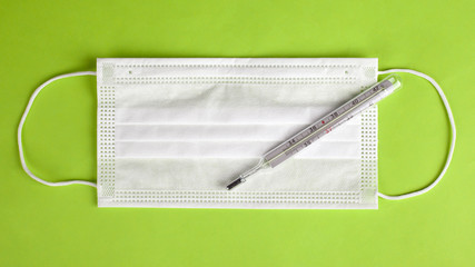 medical face mask with thermometer on a light green background top view. Coronavirus flu protection and screening, quarantine pandemic covid-19