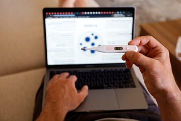 Close up shot of adult man working at home due to coronavirus quarantine concept. Male sitting on couch with laptop, measuring body temperature via digital thermometer. Background, copy space.
