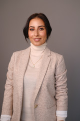 Portrait of a smiling mid-aged business woman in a light milk-colored trouser suit and turtleneck against a light wall.