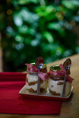 Delicious Italian tiramisu for Christmas table in glass jars decorated with a pink ribbon with a silver spoon, on a red napkin against the background of lights and bokeh.