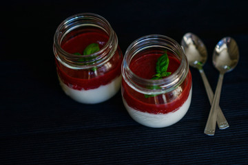 Tasty fragrant panacotta with strawberry compote decorated with fresh basil in glass jars next to silver spoons on a black background. View from above.