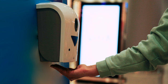 Macro shot of young woman using automatic hand sanitizer dispenser at the international airport with blank glowing advertisement banner on background. Close up, copy space.