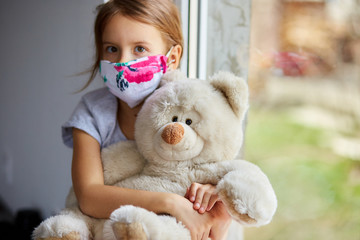Little girl, child in mask with teddy bear sits on windows, coronavirus quarantine - obrazy, fototapety, plakaty