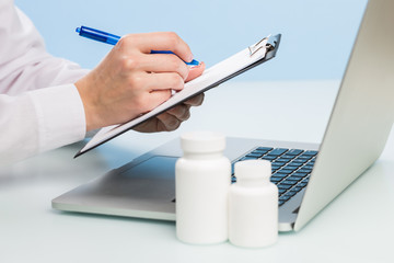 Doctor's hand uses a laptop and writes with a pen on paper on a blue background