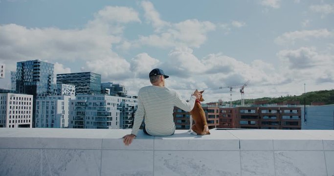 Hipster man and pet brown dog sit on edge of rooftop. Guy and best friend animal look over on empty city on sunny summer day. Social isolation concept. Peace and tranquility in middle of city buzz