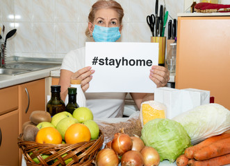 Mature woman in medical mask sitting at the kitchen shows a banner with Stay at home - social media campaign for coronavirus prevention. Quarantine, advice to stop COVID-19 spreading. Global pandemic.