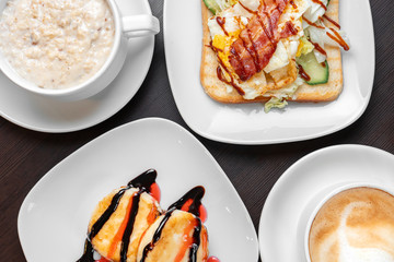 Breakfast consisting of oatmeal porridge with cheesecakes and cappuccino with sandwich, top view, close-up.