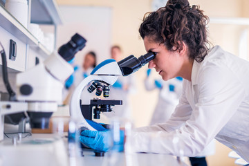 Young female scientist looking through a microscope in a laboratory doing research on finding medicine pharmacy cure vaccine  - obrazy, fototapety, plakaty