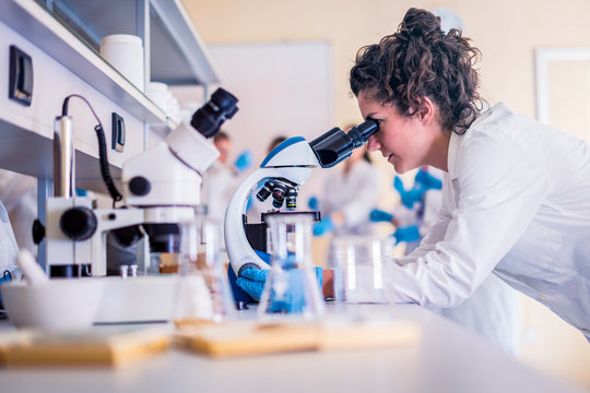 Young female scientist looking through a microscope in a laboratory doing research on finding medicine pharmacy cure vaccine 