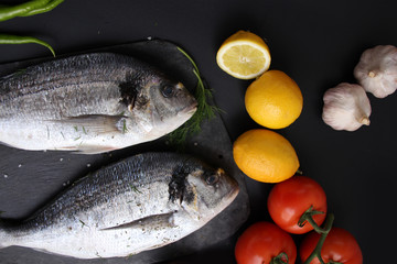 lemons, tomatoes, hot peppers and two carcasses fresh raw atlantic saltwater fish Sparus aurata with shiny silver scales lie on a black background, cooking concept
