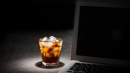 glass of cola with ice and laptop on grey stone table on black background banner