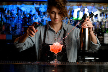 woman bartender expressive pouring cocktail from steel siphon to glass on bar counter.