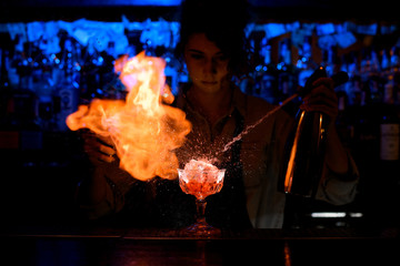 woman bartender expressive pouring cocktail from steel siphon to glass on bar counter.