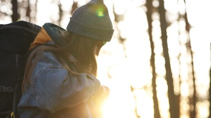 Young charming attractive girl in jeans clothes and a gray knitted hat fastens fastenings on a hiking backpack in the forest in sunny weather. Walk through the woods on a nice spring day. Sunset light - Powered by Adobe
