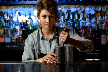 Young handsome woman barman in black apron holds steel glass and metal madler.