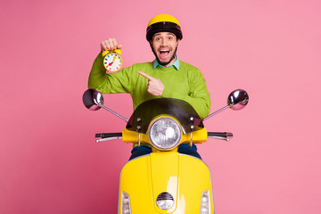 Portrait of his he nice attractive glad cheerful cheery guy sitting on moped holding in hand demonstrating recommending funny alarm clock isolated over pink pastel color background