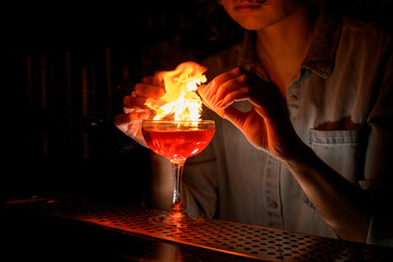 Professional woman barman carefully sprinkles citrus juice on wineglass with cocktail at bar and make fire.