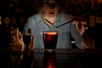Woman barman gently decorates red cocktail with spikelet using tweezers