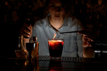 Woman barman decorates red cocktail, sprinkles on it and makes fire.