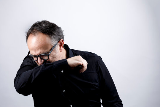 uomo in camicia nera con occhiali starnutisce e si ripara con il gomito, isolato su sfondo nero