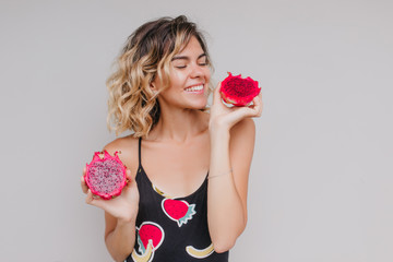 Studio shot of cheerful short-haired girl looking at juicy pitaya. Indoor photo of spectacular tanned woman isolated on light background with dragon fruit.