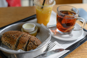 On a tray lies freshly baked sliced bread with seeds, next to butter and sauce. Black tea with lemon and freshly orange juice with a straw in a disposable glass. Breakfast or meal in the cafeteria.